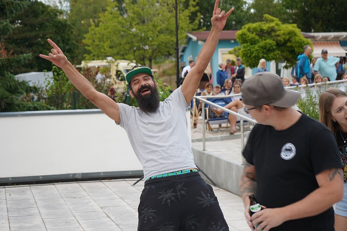 Ein lachender Mann mit Cap und Bart reißt auf dem Festival die Arme hoch, neben ihm weitere fröhliche Besucher in lockerer Stimmung.