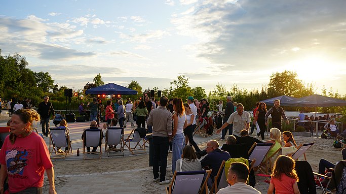 Besucher sitzen in Liegestühlen und stehen vor einer Open-Air-Bühne beim Rock-am-Beach-Event am Eckert Beach bei Sonnenuntergang