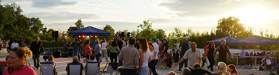 Besucher sitzen in Liegestühlen und stehen vor einer Open-Air-Bühne beim Rock-am-Beach-Event am Eckert Beach bei Sonnenuntergang
