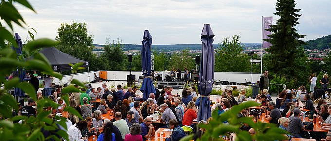 Ein großer Biergarten mit vielen Besucherinnen und Besuchern vor einer kleinen Bühne im Hintergrund. Im Vordergrund rahmen grüne Blätter das Bild ein, im Hintergrund öffnet sich der Blick ins Umland von Regenstauf.