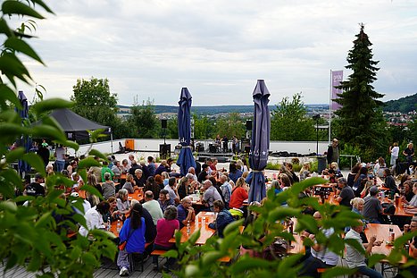 Ein großer Biergarten mit vielen Besucherinnen und Besuchern vor einer kleinen Bühne im Hintergrund. Im Vordergrund rahmen grüne Blätter das Bild ein, im Hintergrund öffnet sich der Blick ins Umland von Regenstauf.