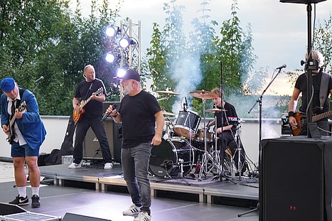 Band mit Sänger, zwei Gitarristen, Bassist und Schlagzeuger bei energiegeladener Live-Performance auf der Bühne mit Lichteffekten und Nebel.