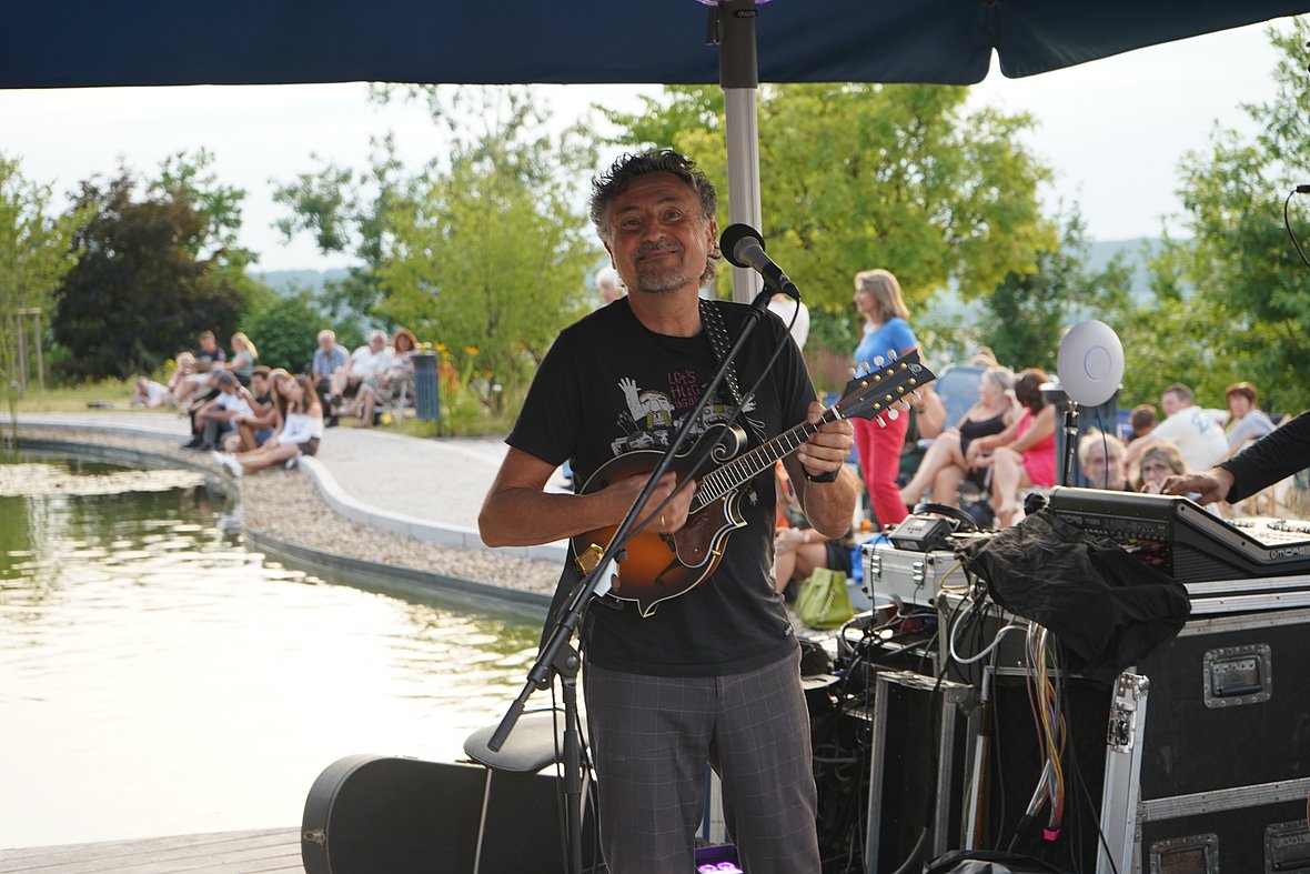 Ein Mann mit dunklem Lockenhaar und schwarzem T-Shirt steht auf einer Bühne am Wasser und spielt eine Mandoline. Er lächelt in die Kamera. Im Hintergrund sitzen viele Menschen auf Steinstufen und am Teichufer.
