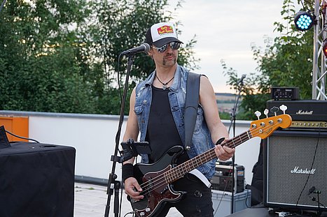 Ein Bassist mit Sonnenbrille, Truckercap und Jeansweste spielt auf der Bühne, im Hintergrund Verstärker und Bühnenlicht.