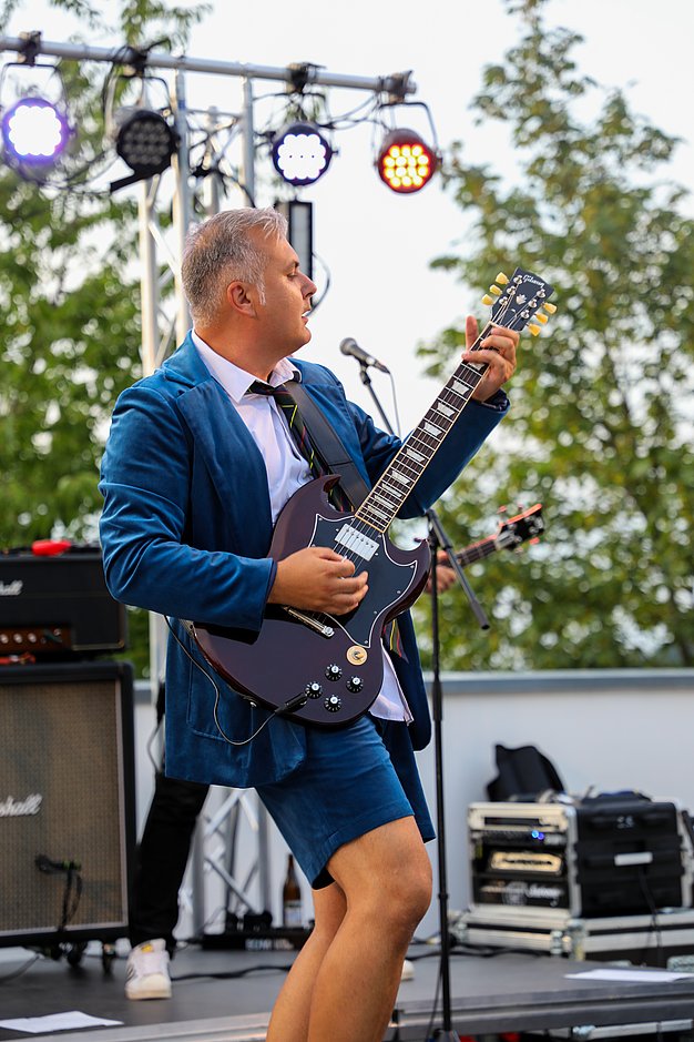 Die Bilder zeigen Eindrücke der Rock-Nacht am Eckert Beach. Zu sehen sind die Live-Auftritte der Bands Rusty Road Runners und ACBarbingCity´25, zahlreiche Besucherinnen und Besucher vor der Bühne sowie die sommerliche Atmosphäre am Strand. In beiden Bands stehen auch Dozenten der Eckert Schulen auf der Bühne. Die Aufnahmen zeigen Musiker, Publikum und die stimmungsvolle Beleuchtung während des Open-Air-Konzerts.