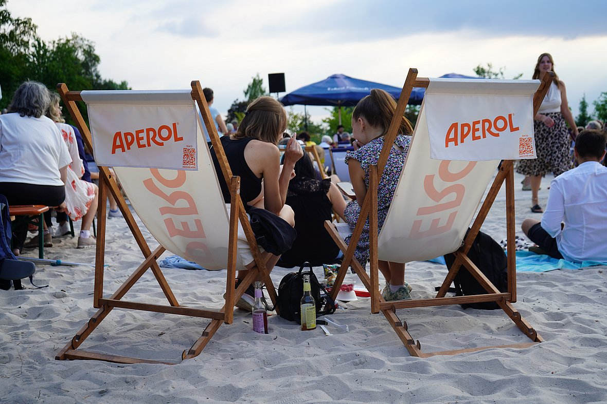 Zwei Frauen sitzen in weißen Liegestühlen mit Aperol-Logo im Sand. Zwischen ihnen stehen bunte Getränkeflaschen. Weitere Menschen sitzen auf Bänken oder auf Picknickdecken, während im Hintergrund blaue Sonnenschirme mit "Eckert Schulen"-Aufdruck und Zuschauer das sommerliche Event genießen.