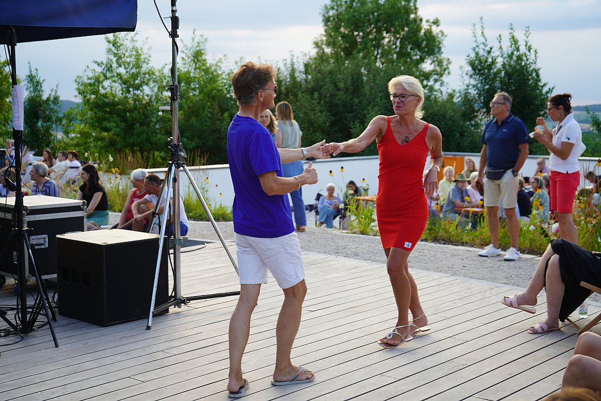 Eine Frau im roten Kleid tanzt mit einem Mann in blauem T-Shirt und weißen Shorts auf einem Holzsteg. Um sie herum sitzen und stehen Menschen in sommerlicher Kleidung, während im Hintergrund Blumenbeete und Bäume den Platz säumen.