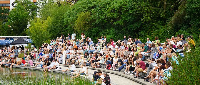 Eine große Menschenmenge sitzt auf terrassenförmig angelegten Steinstufen entlang des Wassers. Einige haben die Füße im Wasser, andere unterhalten sich oder beobachten das Geschehen. Im Vordergrund wachsen Schilfpflanzen, im Hintergrund dichter grüner Bewuchs.
