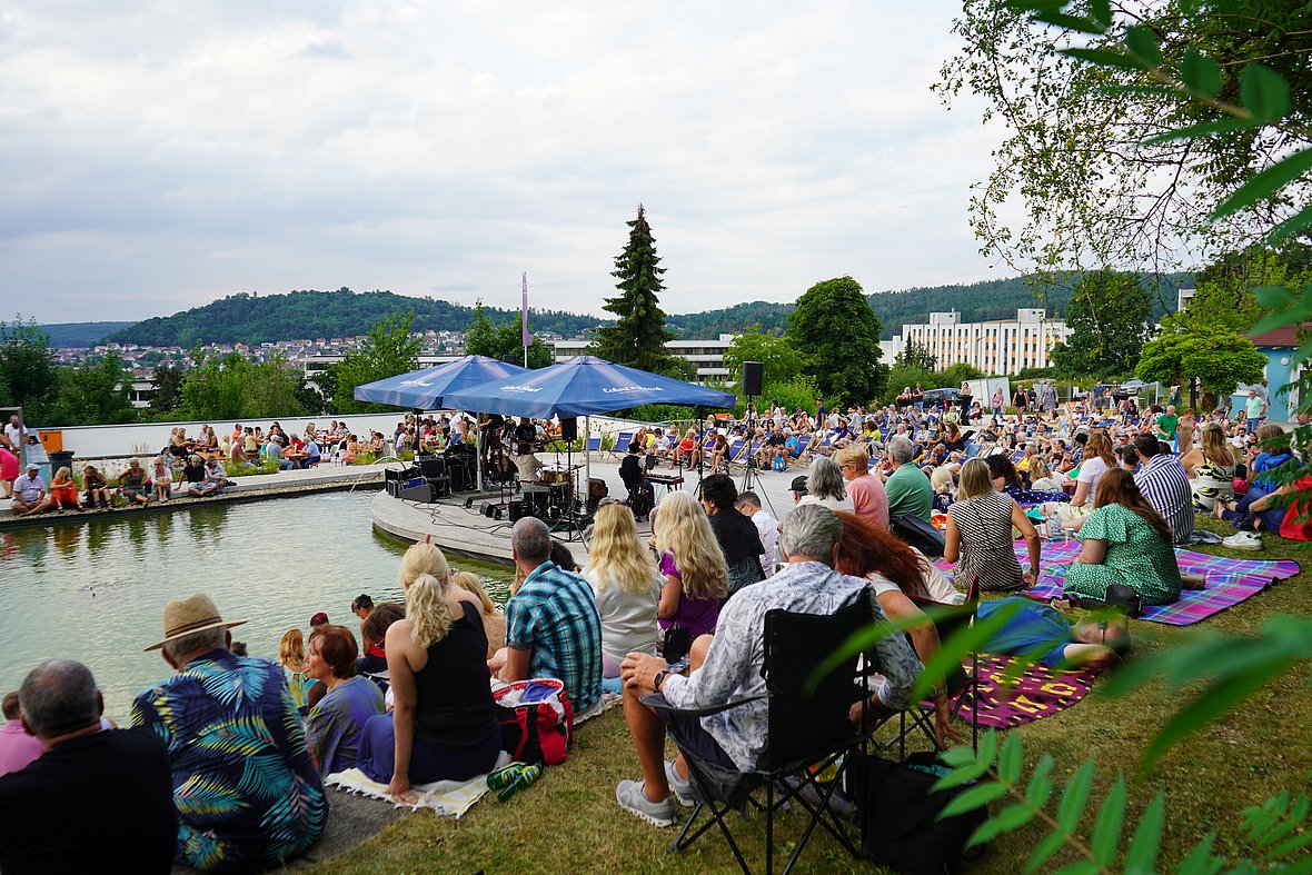 Viele Menschen sitzen auf einer Wiese und rund um einen Teich, einige auf bunten Picknickdecken, andere auf Stühlen. Im Vordergrund steht eine Bühne mit Instrumenten unter zwei blauen Pavillons. Im Hintergrund erstreckt sich ein weiter Blick über die Stadt und die bewaldeten Hügel.