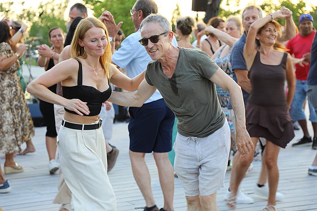 Die Diashow zeigt Eindrücke vom Salsa-Abend am Eckert Beach. Zu sehen sind tanzende Paare, Besucherinnen und Besucher im Publikum sowie die stimmungsvolle Atmosphäre der Veranstaltung am Abend.