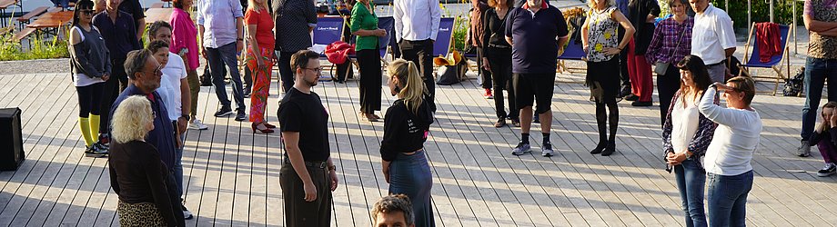 Gruppe von Teilnehmenden steht auf einer Holzplattform unter freiem Himmel bei einem Tanzworkshop im Sonnenuntergang.