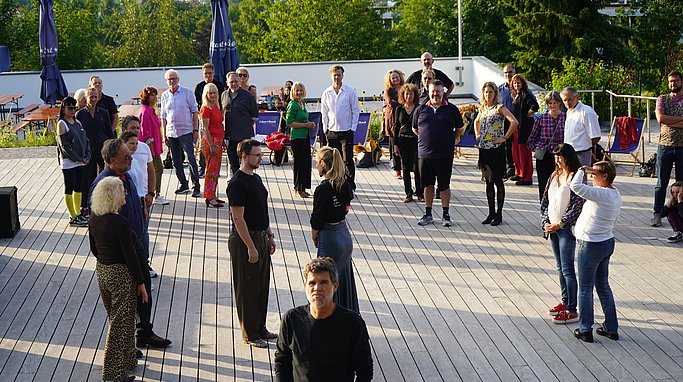 Gruppe von Teilnehmenden steht auf einer Holzplattform unter freiem Himmel bei einem Tanzworkshop im Sonnenuntergang.