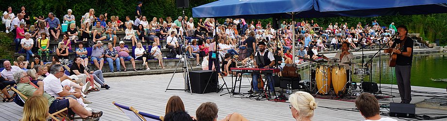 Eine Band spielt auf einer Bühne am Wasser. Zu sehen sind Musiker mit Keyboard, Percussion und Gitarre unter blauen Sonnenschirmen. Viele Menschen sitzen auf Liegestühlen oder den Steinstufen des Amphitheaters, das dicht mit Publikum gefüllt ist. Im Hintergrund dichter Wald.