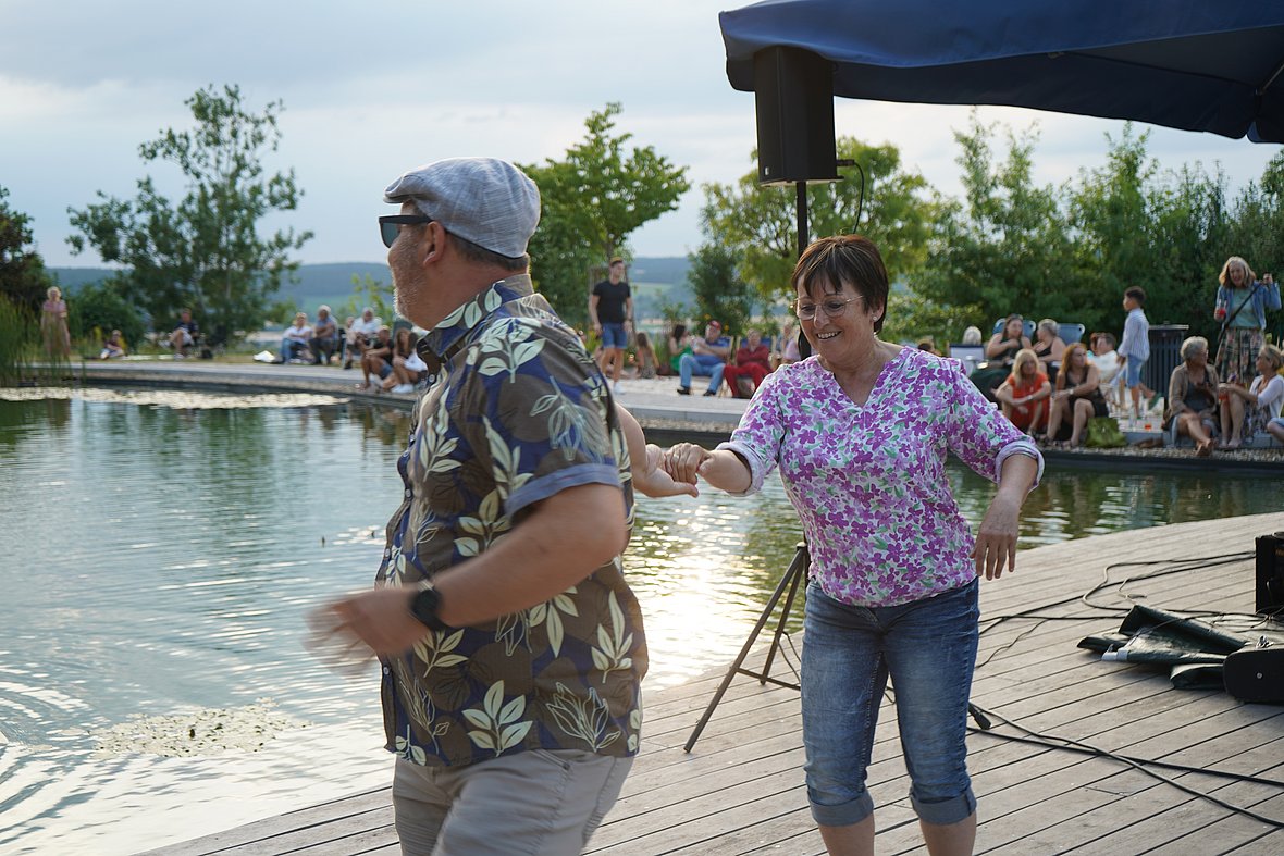 Eine Frau in Jeans und lila Blumenbluse tanzt lachend mit einem Mann im Hawaiihemd und Schirmmütze auf einem Holzsteg an einem Teich. Im Hintergrund sitzen Zuschauer entspannt am Ufer, umgeben von sommerlichem Grün.