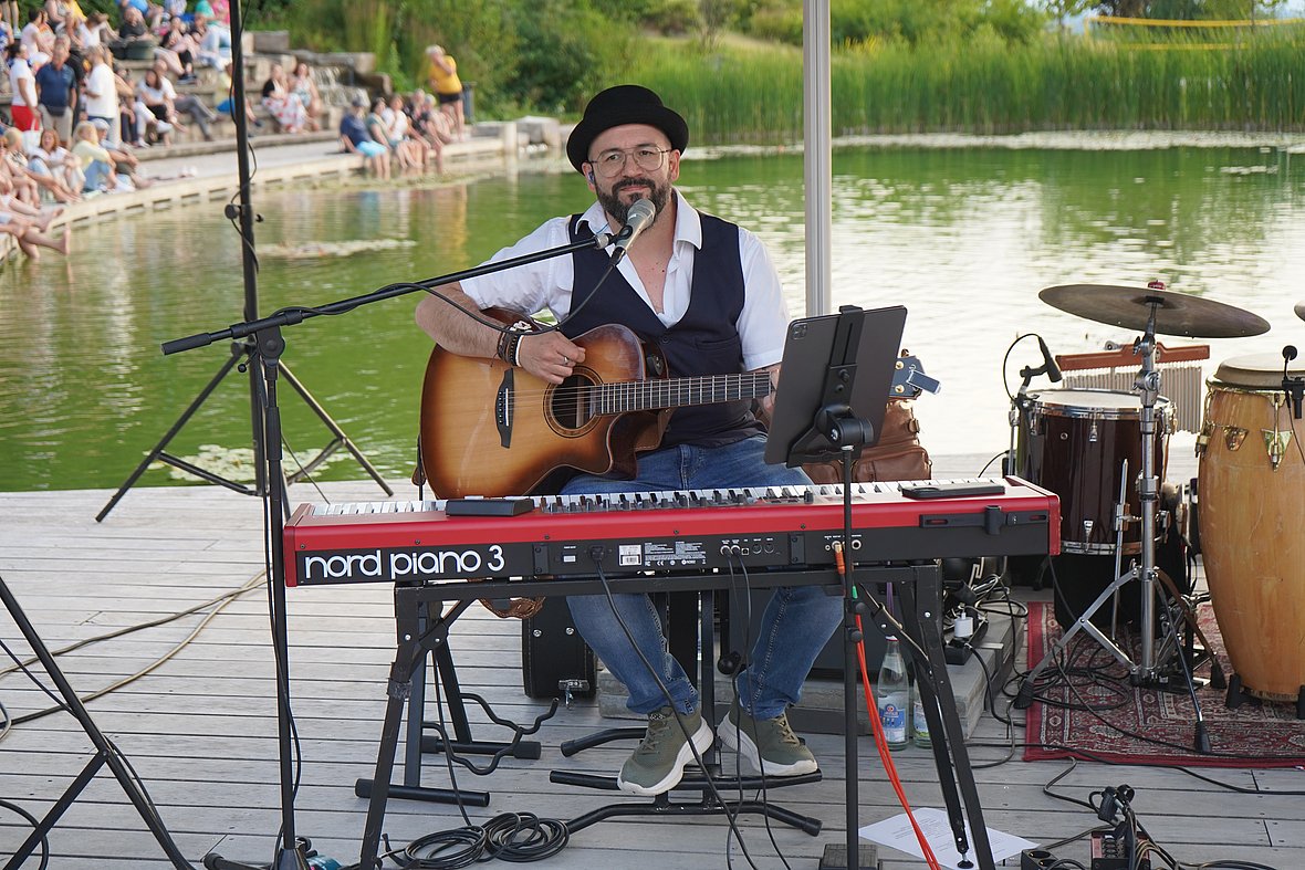 Ein Mann mit schwarzem Hut, Brille und Weste spielt Gitarre und sitzt an einem roten E-Piano auf einer Bühne am Wasser. Hinter ihm sind Schlaginstrumente zu sehen, im Hintergrund sitzen viele Menschen auf den Seeterrassen.