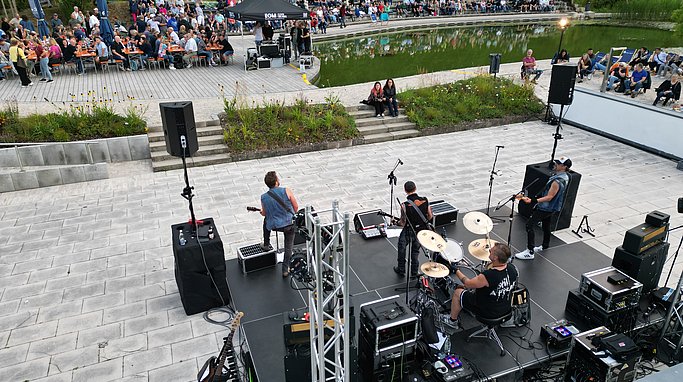 Luftaufnahme eines Open-Air-Konzerts: Eine Rockband spielt auf einer Bühne vor zahlreichen Zuschauerinnen und Zuschauern am Ufer eines Teichs, umgeben von Grünflächen und Tribünen.