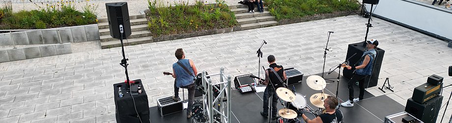 Luftaufnahme eines Open-Air-Konzerts: Eine Rockband spielt auf einer Bühne vor zahlreichen Zuschauerinnen und Zuschauern am Ufer eines Teichs, umgeben von Grünflächen und Tribünen.