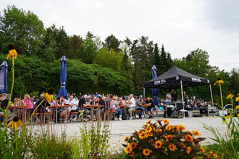 Zahlreiche Menschen sitzen an Bierzeltgarnituren vor einer Bühne am Wasser. Im Vordergrund blühen gelbe und orangefarbene Blumen, rechts steht ein schwarzes Technikzelt mit der Aufschrift „EOM Eventservice“.