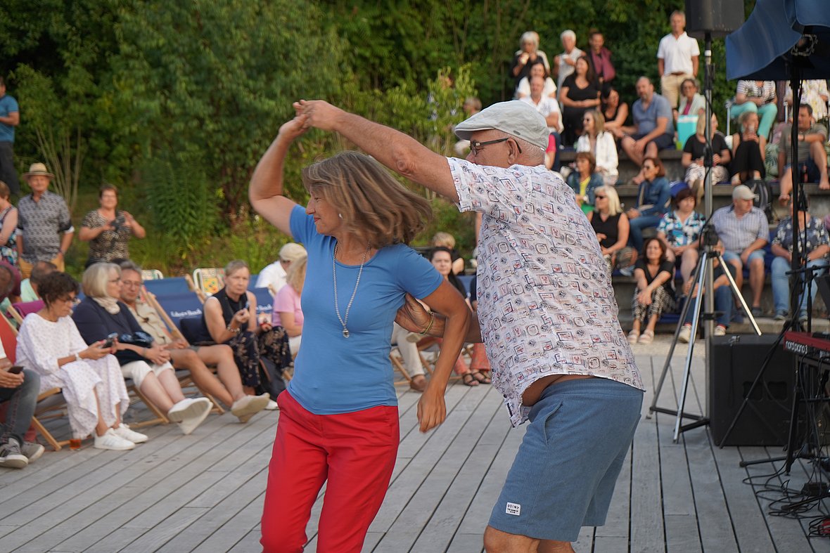 Ein älteres Paar tanzt lachend auf einem Holzsteg vor einem Publikum im Freien. Die Frau trägt ein blaues Shirt und rote Hose, der Mann ein weißes, gemustertes Hemd und eine helle Kappe. Im Hintergrund sitzen viele Menschen auf Stufen.
