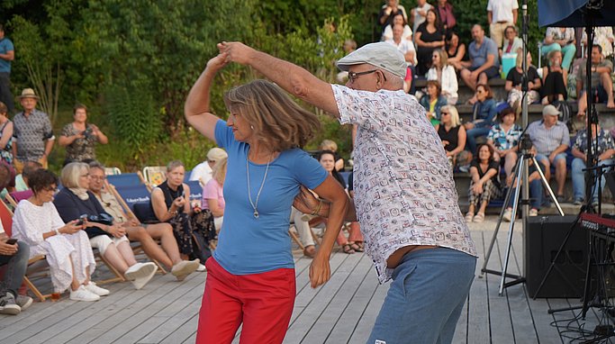 Ein älteres Paar tanzt lachend auf einem Holzsteg vor einem Publikum im Freien. Die Frau trägt ein blaues Shirt und rote Hose, der Mann ein weißes, gemustertes Hemd und eine helle Kappe. Im Hintergrund sitzen viele Menschen auf Stufen.