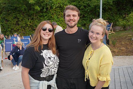 Drei junge Erwachsene stehen Arm in Arm am Eckert-Teich, lachen in die Kamera und zeigen die „Rockhand“. Im Hintergrund sind Zuschauer auf blauen Eckert-Schulen-Liegestühlen zu sehen.