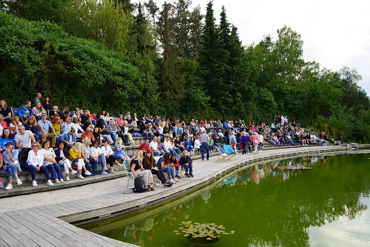 Zuschauer sitzen dicht gedrängt auf den Steinstufen am Teich der Eckert Schulen, während im Vordergrund das grüne Wasser und eine ruhige, sommerliche Stimmung zu sehen sind.
